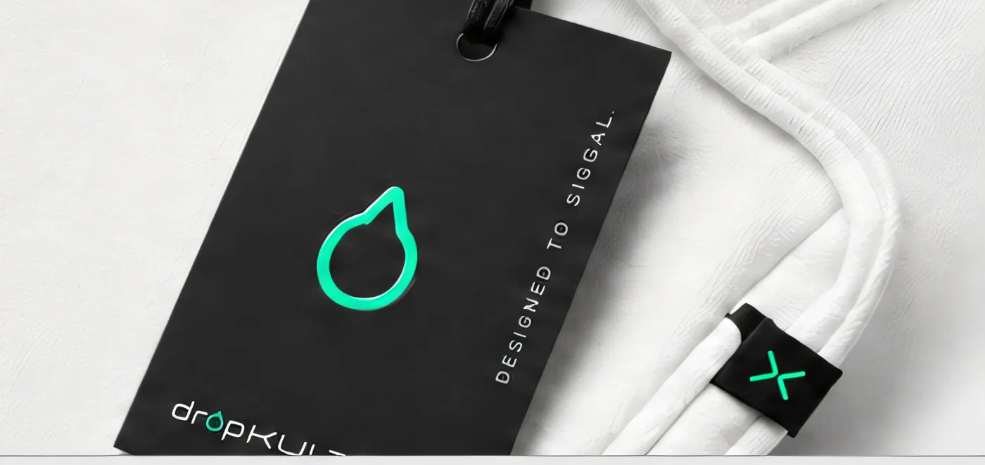 A black dropKULTXR hangtag on a cord against white apparel, printed DESIGNED TO SIGNAL, beside a small X woven label